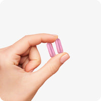 Conceive Plus Women's Combo - Fertility Support + Applicators (IT) featuring a close-up of a hand gently holding two pink vegetarian fertility capsules between fingers against a white background, illustrating capsule size and form for visual clarity.