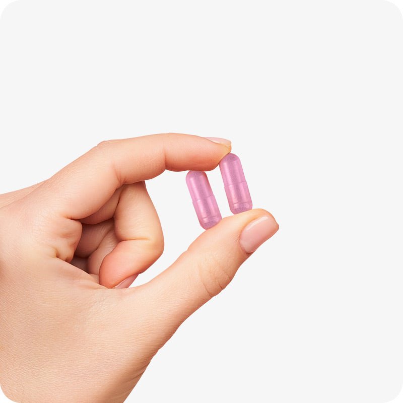 Conceive Plus Women's Combo - Fertility Support + Applicators (IT) featuring a close-up of a hand gently holding two pink vegetarian fertility capsules between fingers against a white background, illustrating capsule size and form for visual clarity.